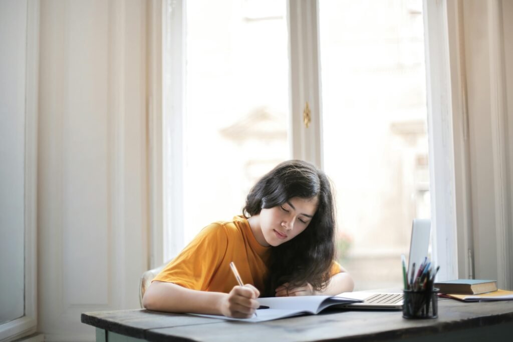 A young woman focused on writing in her notebook near a sunlit window.