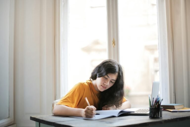 A young woman focused on writing in her notebook near a sunlit window.