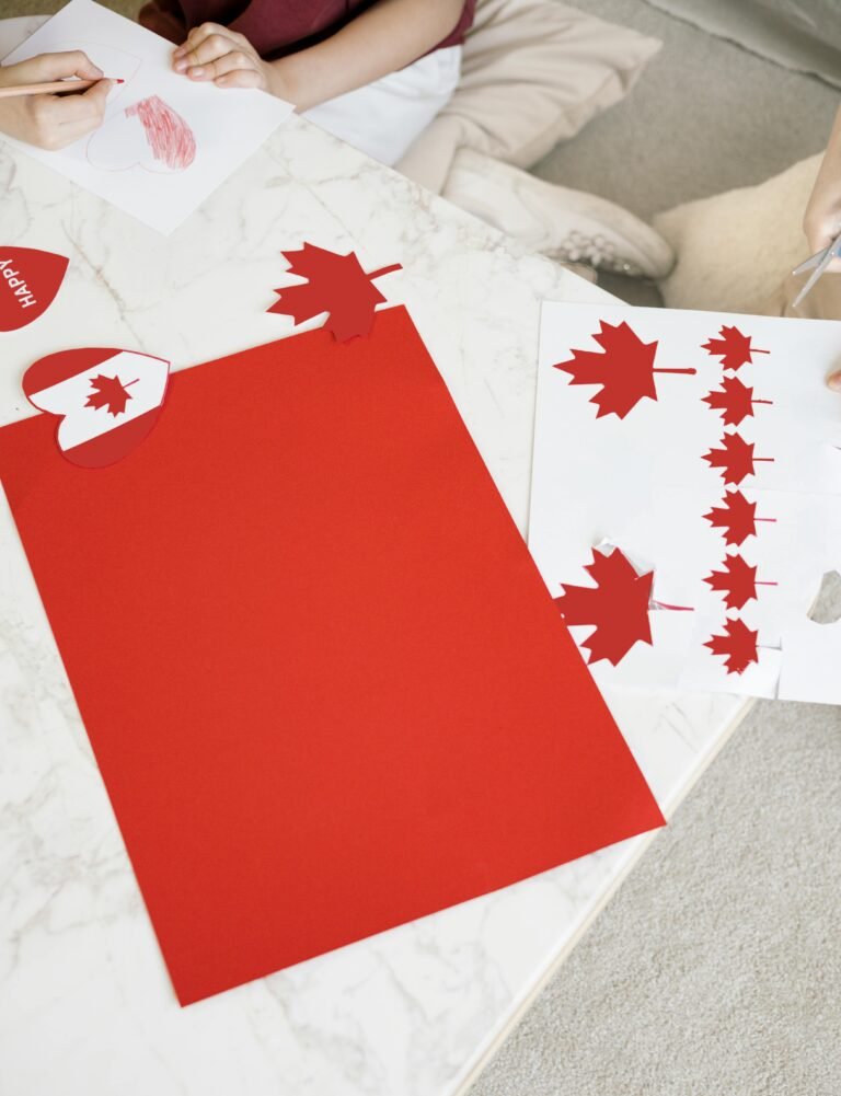 Hands crafting Canada Day decorations with red paper and maple leaves.