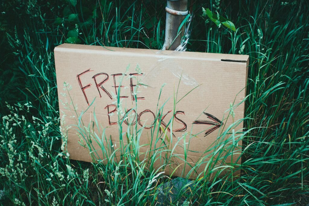 A cardboard sign with 'Free Books' handwritten, placed among green grass outdoors promoting book exchange.
